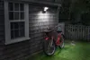 A red bicycle with a basket is parked next to a wooden-shingled house, illuminated by an outdoor spotlight. Produktbild-Vorschau