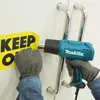 A person wearing gloves is using a heat gun to remove a yellow sticker with black text from a white surface with chrome handles. Podgląd zdjęcia produktu
