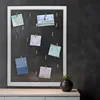 A white framed photo holder with multiple photos clipped to strings, displayed on a dark textured wall above a white cabinet. A small potted plant is on the cabinet to the right. Náhled obrázku produktu