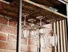 A close-up view of a wine rack mounted under a wooden shelf, holding several wine glasses. The background shows a brick wall and vertical wooden slats. Podgląd zdjęcia produktu