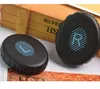 Two black ear pads with blue and white striped oval inserts are placed on a surface with a wooden box in the background. Produktbild-Vorschau