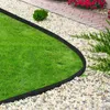 A black flexible garden border separating a lush green lawn from a path made of light-colored gravel and red bricks, with some greenery and red flowers in the background. Produktbild-Vorschau