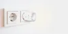 A white smart plug is inserted into a white electrical outlet on a white wall. Voir les images du produit