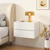 A white floating nightstand with two drawers is shown next to a bed. A lamp, a cup, and a vase with flowers are on and next to the nightstand. Podgląd zdjęcia produktu