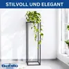 A tall, black, rectangular metal plant stand with a green potted plant on top, placed against a white wall with soft lighting. Produktbild-Vorschau
