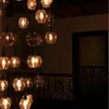 A collection of vintage-style pendant lights hanging in a dimly lit interior, possibly a staircase or hallway, with a dark railing and a window in the background. Podgląd zdjęcia produktu