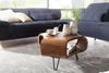 A wooden coffee table with a modern, curved design is placed on a rug in front of a dark blue sectional sofa. Two vases with white flowers are on the table, along with a cup and saucer. Produktbild-Vorschau