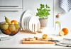 A kitchen counter scene featuring a plate stand with white plates, a fruit bowl, a plant, and a water bottle. Náhled obrázku produktu