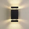 A black rectangular outdoor wall light mounted on a textured wall, emitting light upwards and downwards. Anteprima dell’immagine prodotto