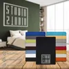 A stack of colorful bed sheets is displayed on a wooden surface in front of a bedroom scene. The sheets are neatly folded, and some have the brand logo visible. Icons indicating product features are also present. Produktbild-Vorschau