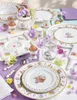 A beautifully set Easter-themed table with plates, egg cups, a watering can vase with flowers, and other decorative items. Produktbild-Vorschau