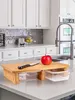 A kitchen counter with a bamboo cutting board with two clear plastic storage containers underneath. A red apple and a knife are on the cutting board. The background shows kitchen cabinets and a sink. Produktbild-Vorschau