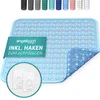 A light blue shower mat with a diamond pattern is shown, partially folded to reveal its underside. A green banner with text and a circular inset showing two white adhesive hooks are also present. Produktbild-Vorschau