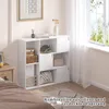 A white cube storage shelf unit with 8 compartments and 3 doors, filled with various items, placed in a bedroom setting next to a window with sheer curtains. Produktbild-Vorschau