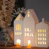 A collection of white ceramic house-shaped tea light holders with illuminated windows, arranged on a wooden surface with dried flowers in a vase in the background. Produktbild-Vorschau