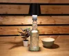 A table lamp made from a glass bottle sits on a wooden table in front of a wooden slatted wall. A small potted plant and a bowl are also on the table. Produktbild-Vorschau