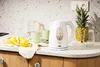 A kitchen counter scene with a white electric kettle, lemons, cups, and a pineapple. Voir les images du produit
