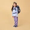 A young girl wearing a backpack stands against a plain background. Náhľad obrázku produktu
