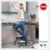 A woman is using a step stool in a kitchen to reach items in a cabinet. Produktbild-Vorschau