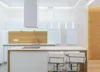 A modern, minimalist kitchen with white cabinets, a wooden backsplash, and a central island with bar stools. Náhled obrázku produktu