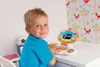 A young boy is sitting at a desk, looking at the camera and smiling, while playing with a toy laptop. The background features colorful wallpaper with a fox and tree pattern. Produktbild-Vorschau