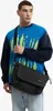 A man wearing a blue and navy sweatshirt and light blue jeans, carrying a black messenger bag across his body. Náhled obrázku produktu