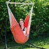 A woman sits in an orange hammock chair outdoors, with lush green foliage in the background. Podgląd zdjęcia produktu