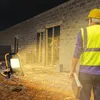 A construction worker in a yellow hard hat and reflective vest stands in front of a building under construction at night, illuminated by a portable LED work light. Produktbild-Vorschau