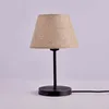 A table lamp with a beige fabric shade and a black metal base and stem, set against a light gray background. Produktbild-Vorschau