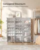 A shoe rack with multiple compartments filled with shoes, bags, and folded clothes, placed in a room with a window and a clothing rack in the background. Produktbild-Vorschau