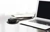 A black wireless earbud charging case sits on a black charging pad next to a stack of papers and a laptop. Produktbild-Vorschau