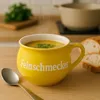 A yellow soup mug with text is placed on a wooden surface, next to sliced bread and a spoon. Steam rises from the soup. Voir les images du produit