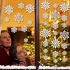 A woman and a girl looking out a window decorated with hanging snowflakes, with a Christmas tree visible in the background. Produktbild-Vorschau