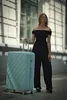 A woman stands next to a large mint green suitcase on a sidewalk. Produktbild-Vorschau