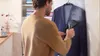 A man is steaming a navy blue blazer with a handheld garment steamer. The blazer is hanging on a hanger on a wall. In the background, a shelf holds folded clothes and decorative items. Náhľad obrázku produktu
