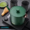 A green electric kettle with a gooseneck spout is placed on a black heating base. Nearby are a matcha whisk and a small dish of matcha powder, along with two dark cups. The scene suggests preparation for making tea. Podgląd zdjęcia produktu
