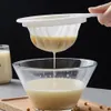 A handheld strainer is used to filter a light-colored liquid into a glass bowl. Produktbild-Vorschau