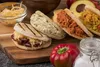 A close-up shot of several arepas filled with various ingredients, arranged on a wooden board with some fresh vegetables and a jar of salt in the foreground. Produktbild-Vorschau
