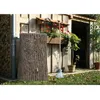 A rain barrel designed to look like a tree trunk stands next to a wooden shed, with plants and a broom nearby. Produktbild-Vorschau
