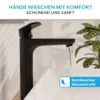 A black faucet with water flowing into a white sink, with a plant in the background. Produktbild-Vorschau