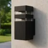 A black outdoor wall light fixture is mounted on a textured concrete wall, with a blurred background of greenery and a pathway. Náhľad obrázku produktu