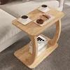 A C-shaped side table with a wood-like finish is placed next to a sofa, holding two cups of coffee and a plate of donuts. Books are stacked on the lower shelf. Náhled obrázku produktu