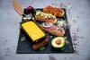 A slate serving board with a raclette grill filled with melted cheese, hot dogs, avocado, and sprouts. Náhled obrázku produktu