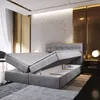 A modern bedroom with a grey upholstered boxspring bed with its storage compartments open. Produktbild-Vorschau