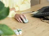 A person's hands using a wireless mouse on a wooden desk, with a laptop and other items in the background. Produktbild-Vorschau