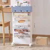 A white and blue rolling storage cart filled with books and magazines, next to a wooden chair and desk. A cardboard box with 'STORAGE RACK' printed on it is visible in the lower right corner. Produktbild-Vorschau