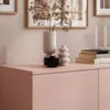 A close-up of a pink dresser with decorative items on top, including a vase with dried flowers, a black bowl, and books. Produktbild-Vorschau