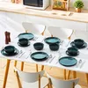 A dining table set with teal and black stoneware dishes, cutlery, and salt and pepper shakers, set against a kitchen counter with a microwave and shelving. Náhled obrázku produktu