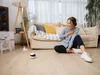 A woman is sitting on the floor in a living room, looking at a small robot. Náhled obrázku produktu