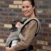 A woman is holding a baby in a carrier in front of a brick wall. Náhľad obrázku produktu